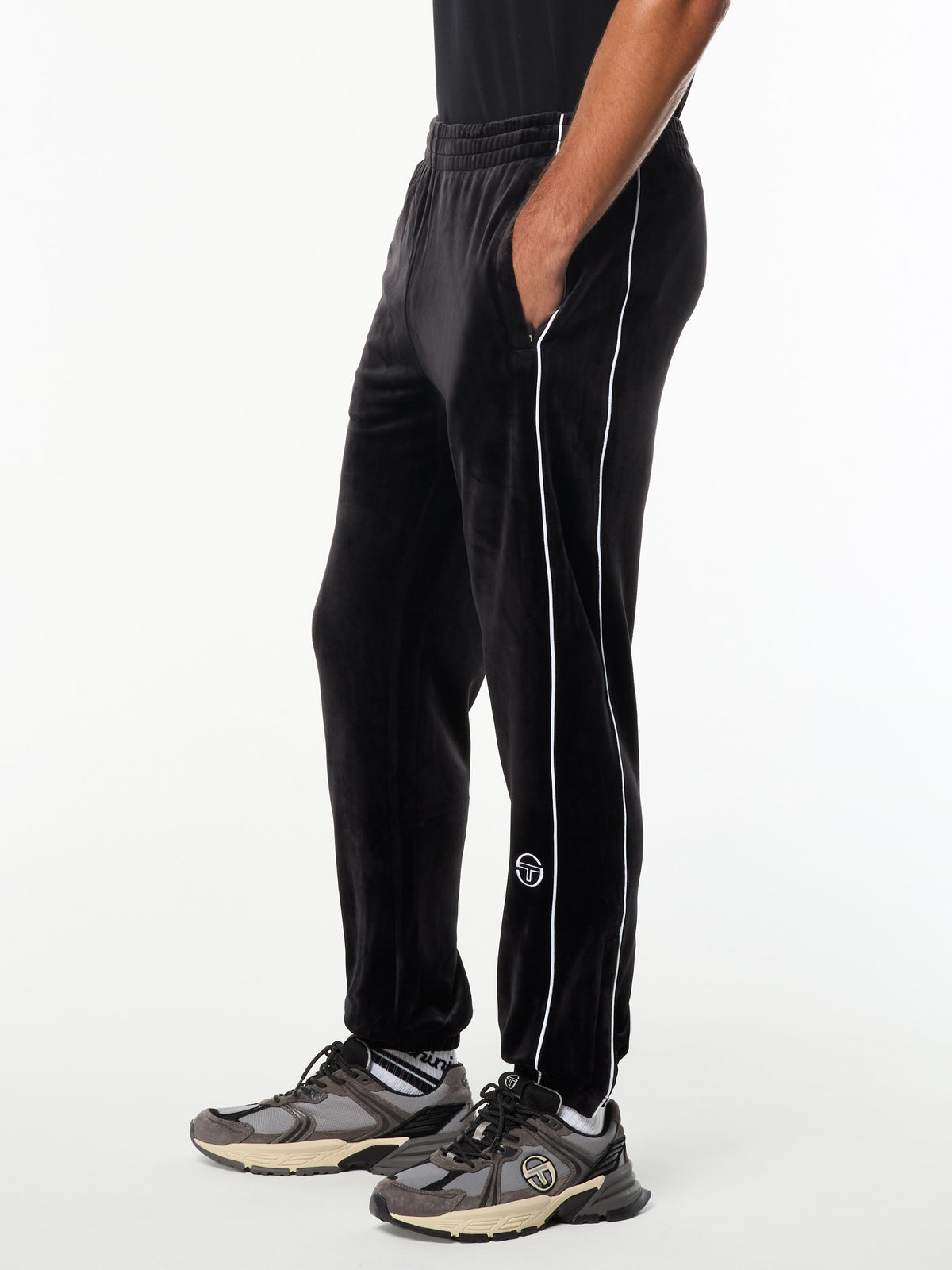 Side view of the Lioni Velour Track Pant in Black Beauty with white piping and embroidered logo, worn with gray sneakers.