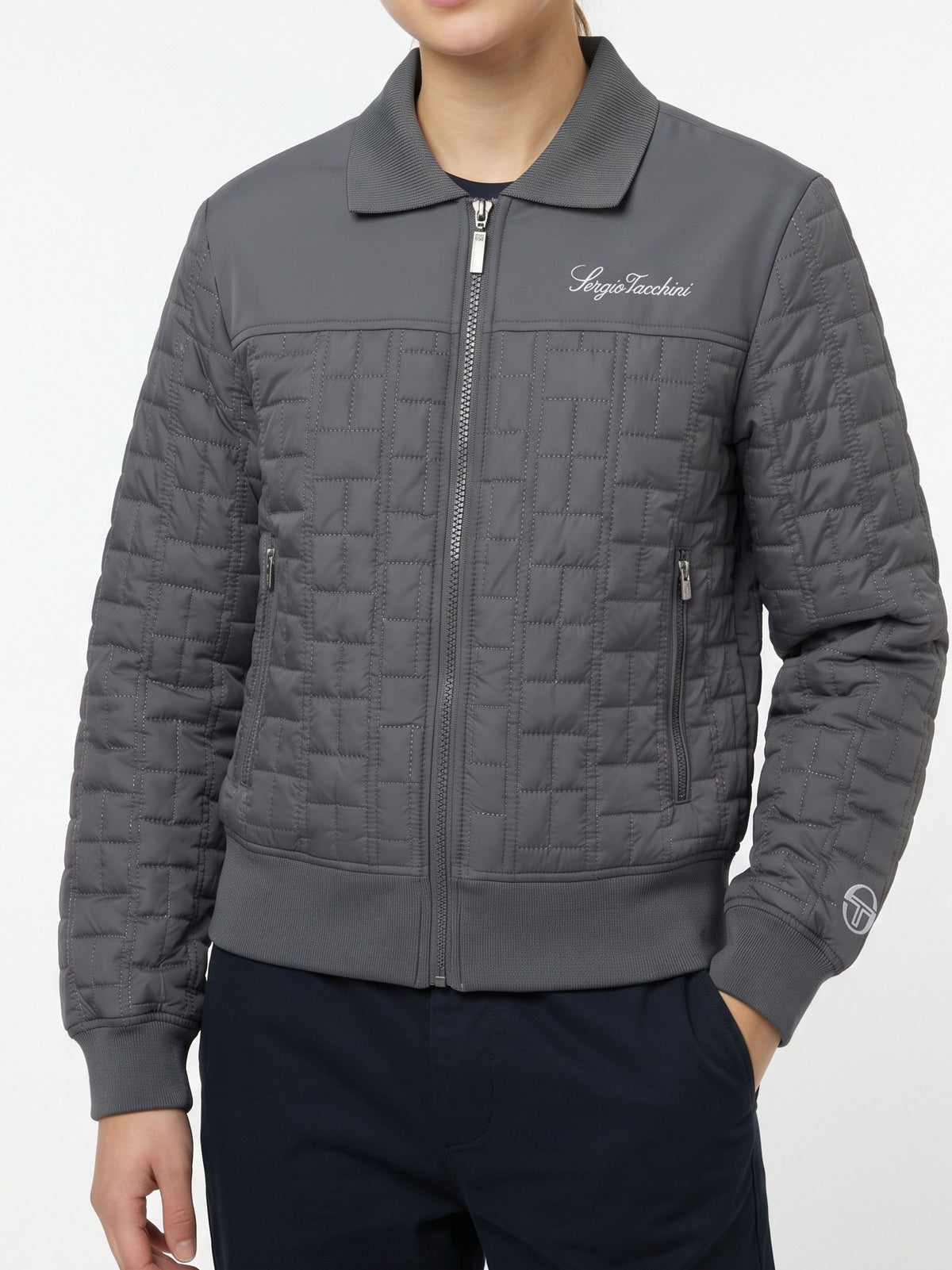 Model wearing the Women's Anji Quilted Track Jacket in Quiet Shade, front view of the grey quilted zip jacket with a smooth upper yoke, script chest embroidery, zip pockets, ribbed collar, cuffs, and hem, and a logo on the sleeve cuff, hand in the pocket of navy pants.