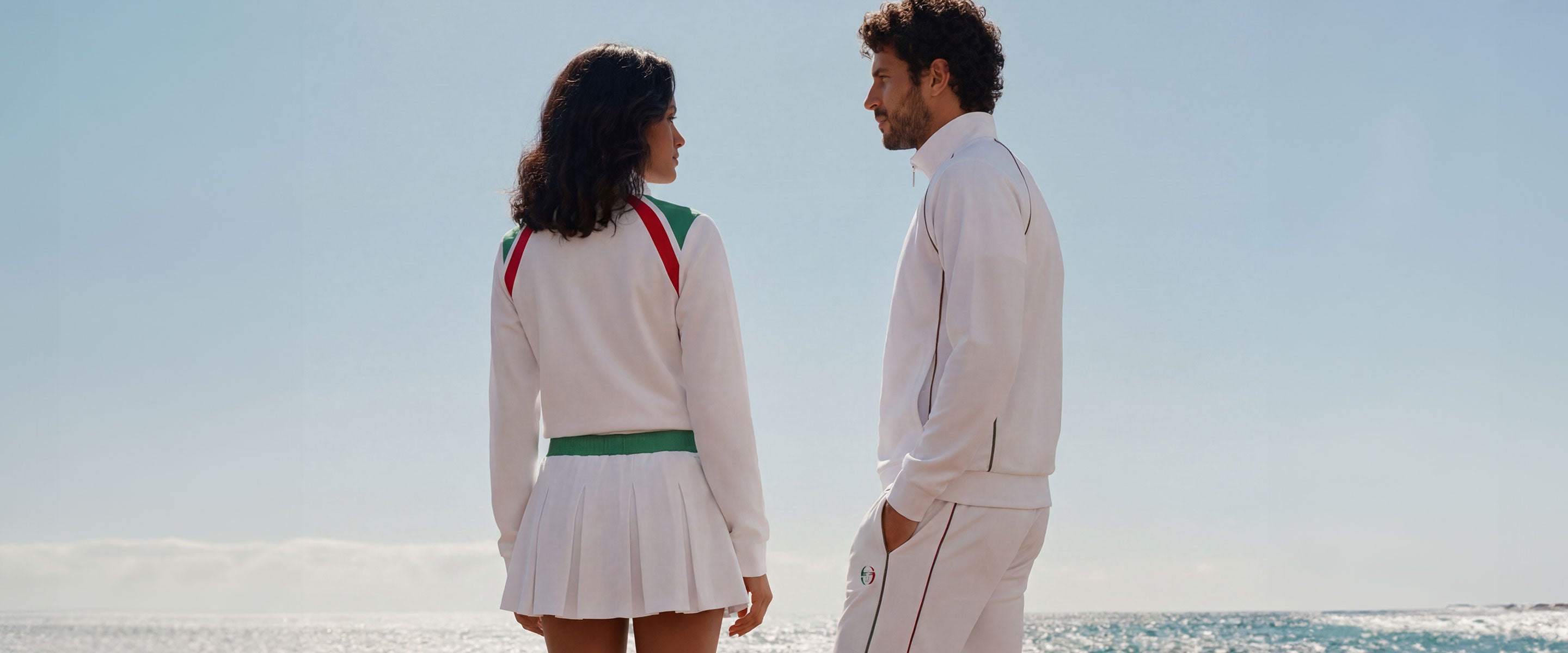 Man and woman standing seaside wearing white sets with red and green accent details.