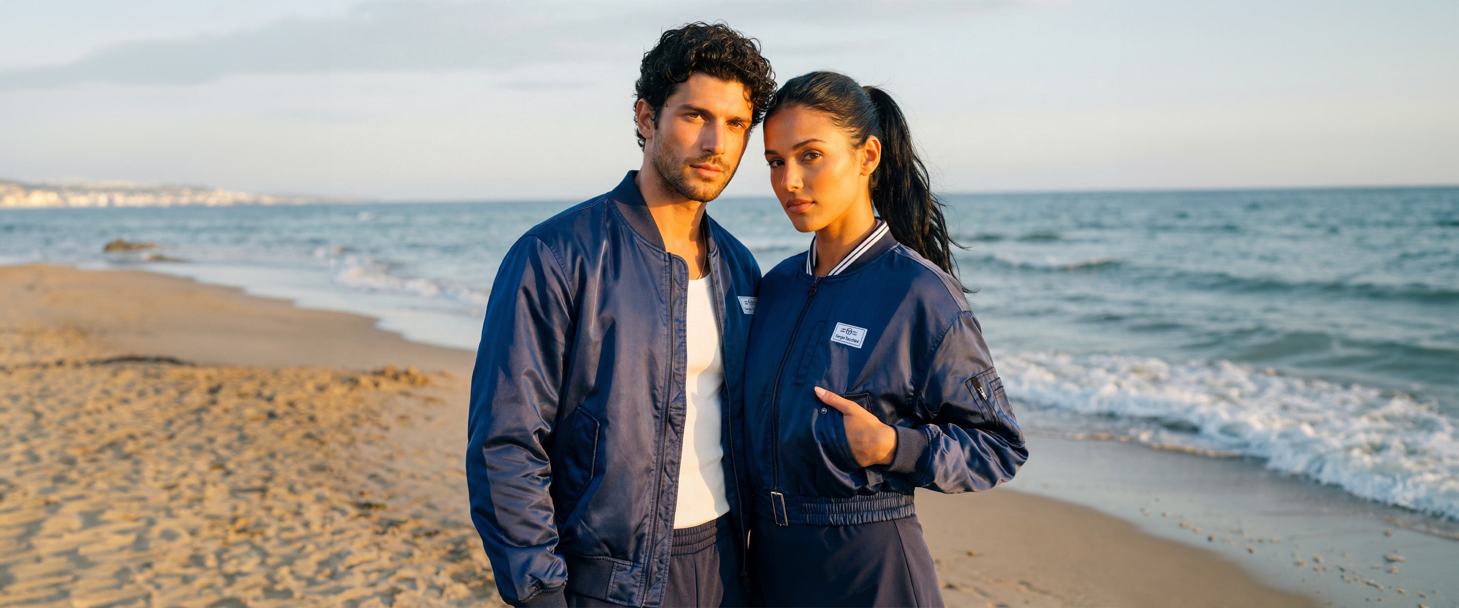 Man and woman standing on a beach wearing matching navy blue bomber jackets. 