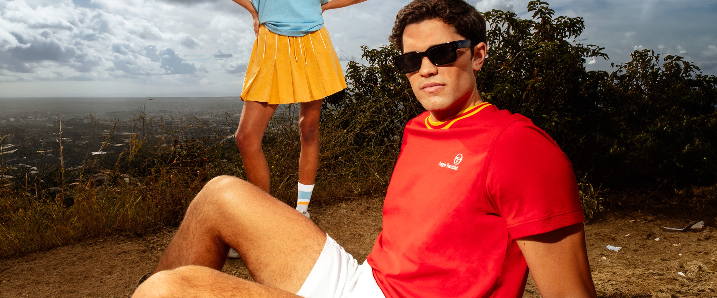Man sitting on ground wearing a red shirt with a woman standing behind him wearing a blue shirt with a yellow skirt.