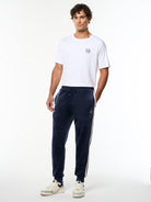 Model wearing the Damarindo Velour Track Pant in Maritime Blue with a white logo tee, full body front view.