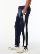 Side view of the Damarindo Velour Track Pant in Maritime Blue with white side stripes, worn with cream sneakers.