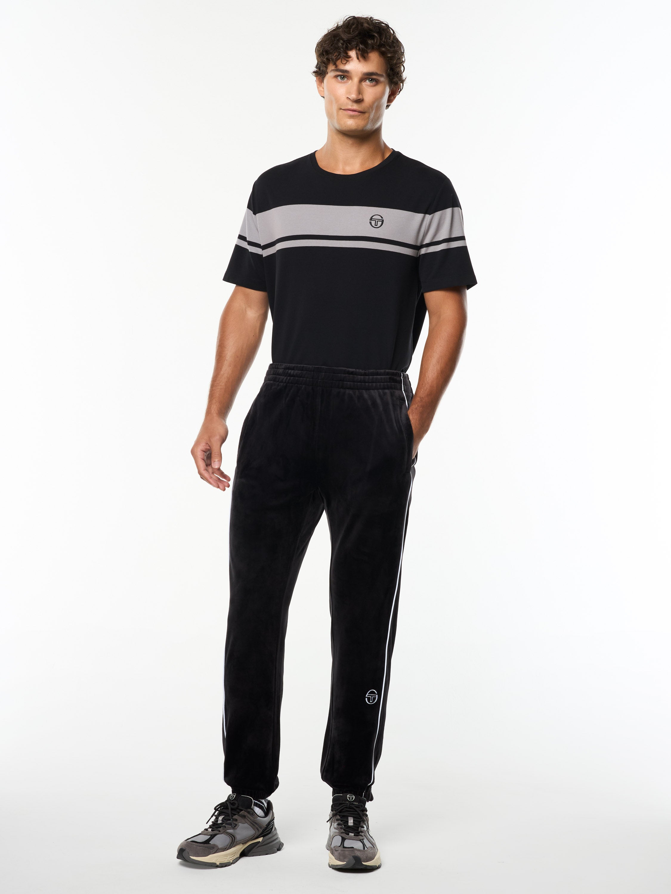 Model wearing the Lioni Velour Track Pant in Black Beauty with a black and gray striped tee, full body front view.