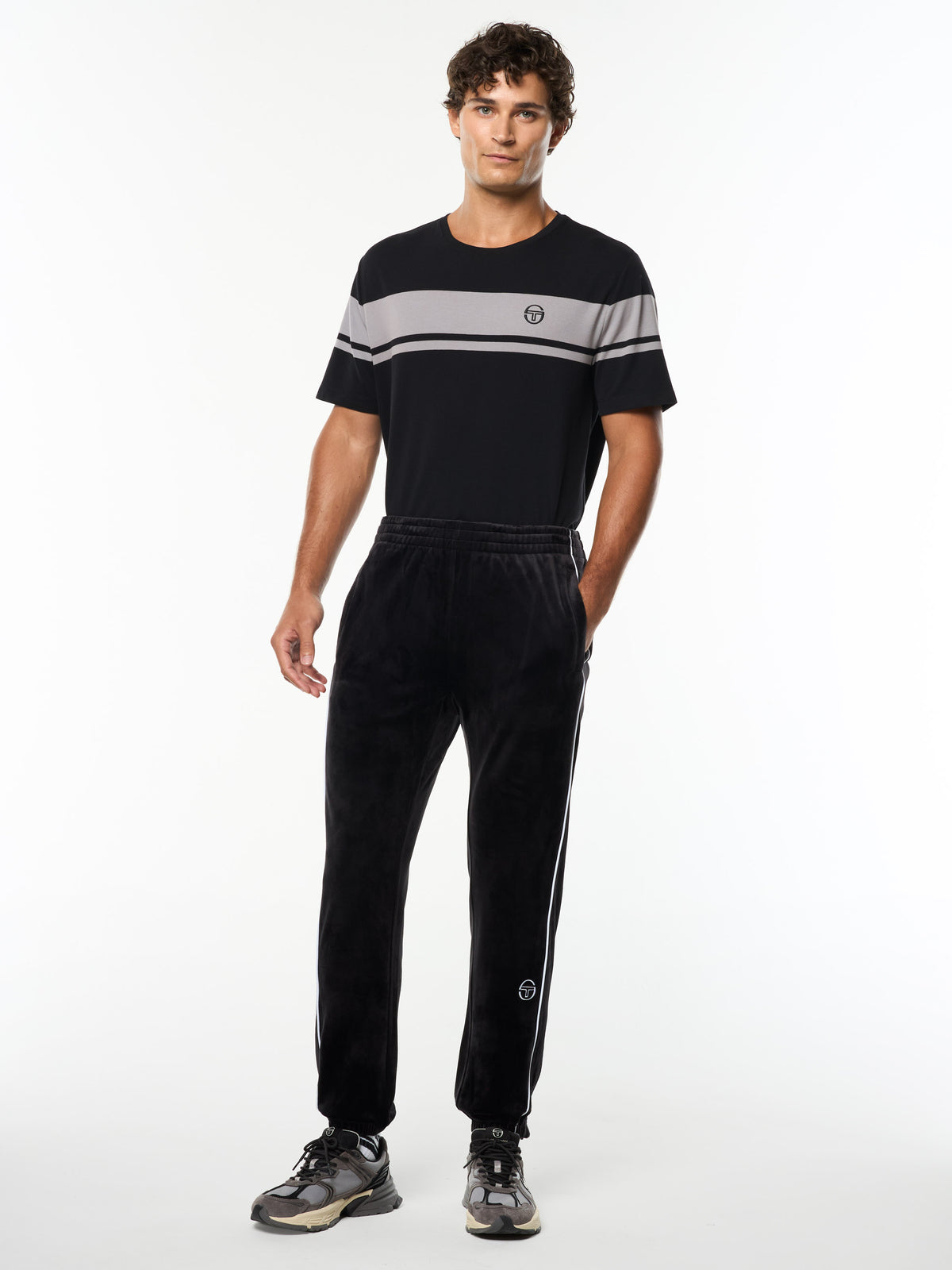 Model wearing the Lioni Velour Track Pant in Black Beauty with a black and gray striped tee, full body front view.