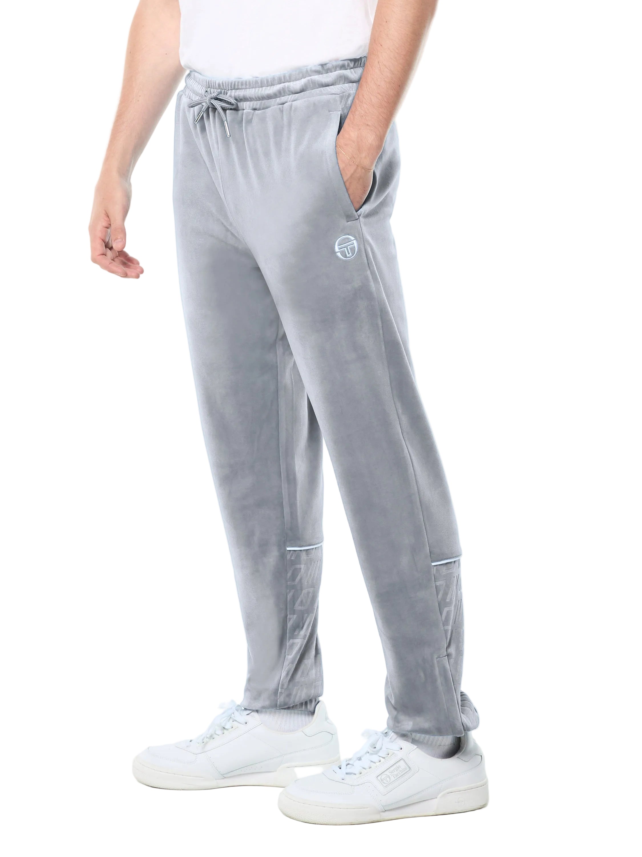 A person wearing light gray velvet jogger pants with a drawstring waist, side pockets, and a small circular logo patch on the upper left thigh, paired with white sneakers against a plain white studio background.